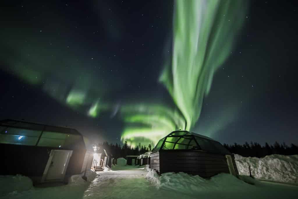 Ground covered in thick snow looking up at the northern lights with buildings in the background