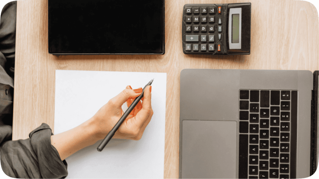 Someone with a pen in their hand above a computer, calculator and iPad on a table